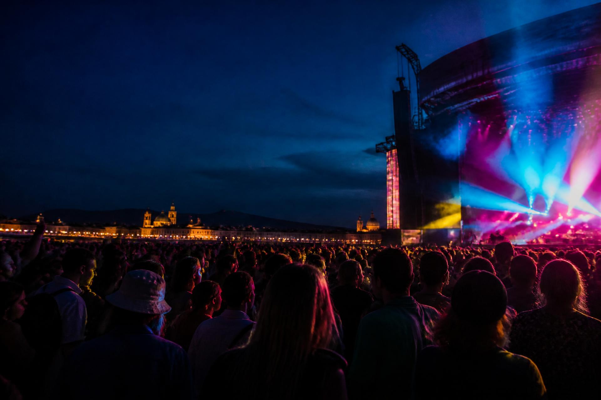 A concert crowd facing a luminous stage with a night city skyline behind the venue